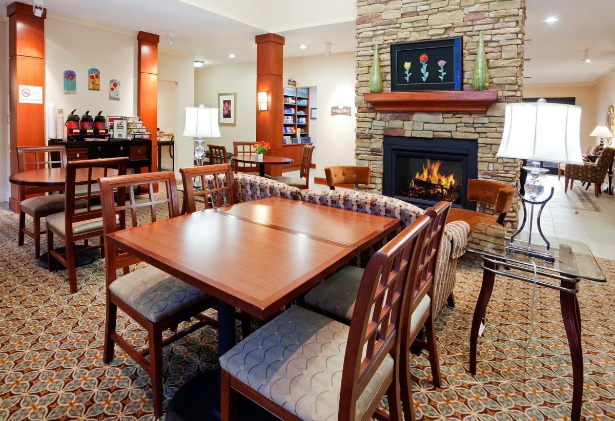 The image presents a warm, communal seating area with a fireplace, multiple dining tables, comfortable chairs, and artistic decor. The environment is ideal for informal gatherings, relaxing by the fire, or enjoying a shared meal—conveying flexibility and a sense of belonging.