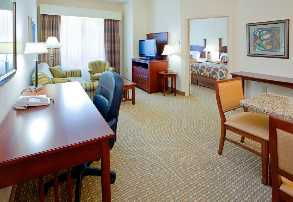 This interior image captures a welcoming, multifunctional living space featuring a dedicated work desk with a comfortable chair, a plush sofa seating area ideal for relaxation, and an open view toward the private bedroom. Natural light brightens the room, and the layout is both practical and inviting, perfect for business or leisure travelers.