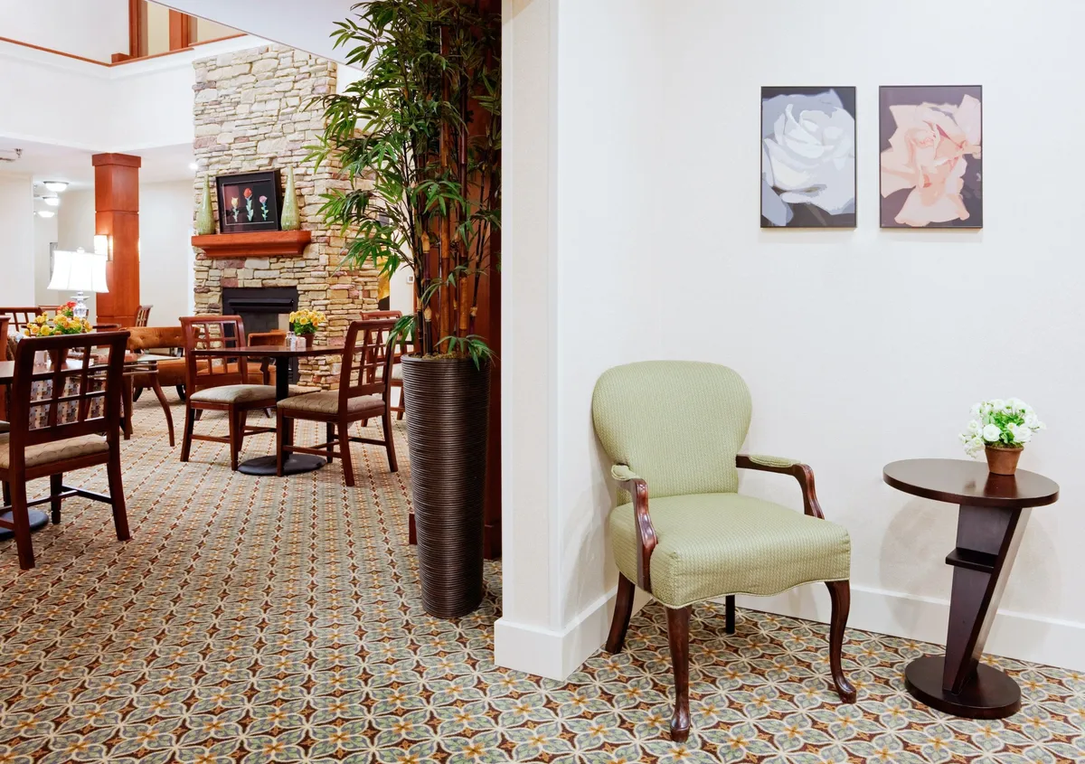 A softly lit, inviting interior corner featuring a green upholstered chair beside a small side table, complemented by vibrant flower paintings and a potted plant. The ambiance feels peaceful and calm, suggesting a quiet spot to relax or read. A fireplace adds a further touch of homeliness and warmth.