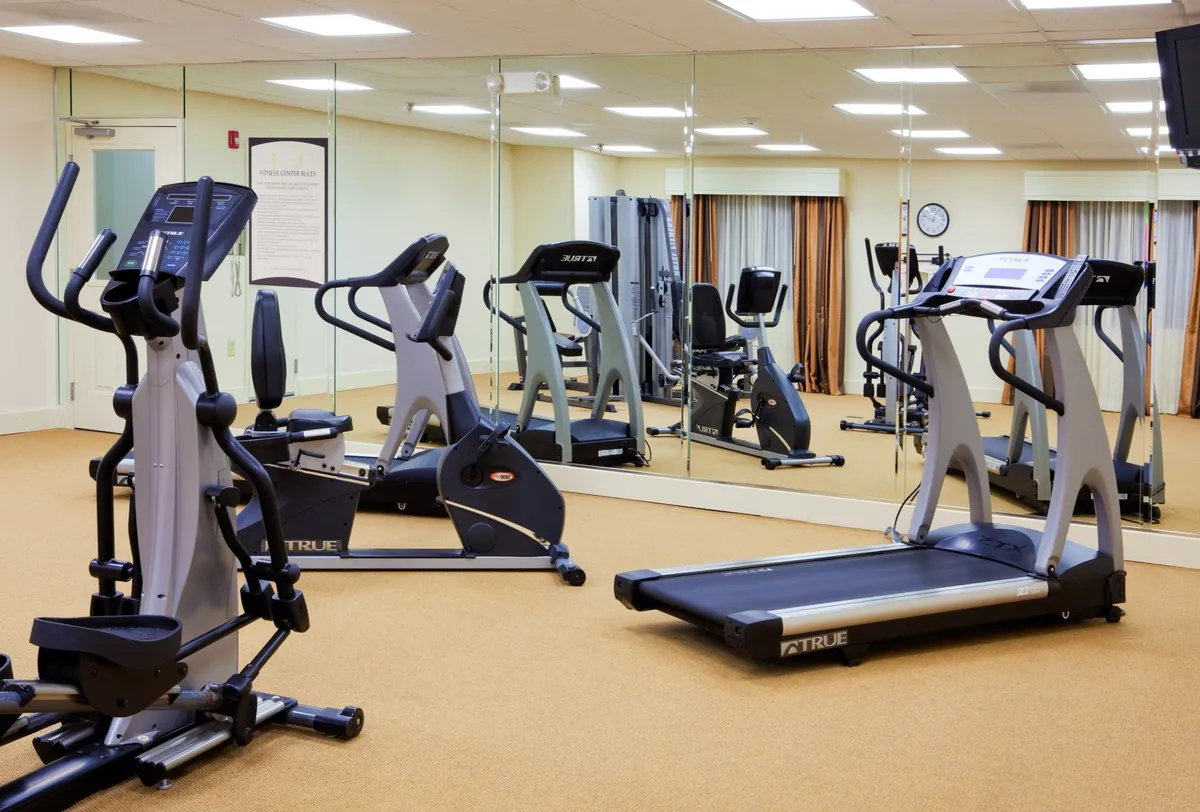 A bright, modern fitness center equipped with an elliptical machine, stationary bike, and treadmill, all set against a mirrored wall that visually expands the space. The energetic ambiance is enhanced by plenty of natural light, providing a motivating environment for workouts.