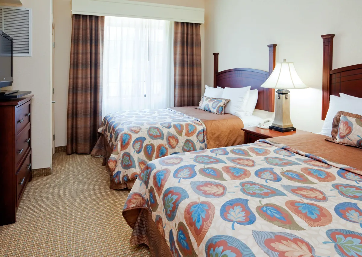 A cozy bedroom scene with two comfortable double beds placed side by side, softly illuminated by natural light streaming in from a window. A bedside lamp offers a warm glow, and the space feels welcoming, perfect for unwinding after a long day. The room subtly hints at both relaxation and togetherness, ideal for families or companions.