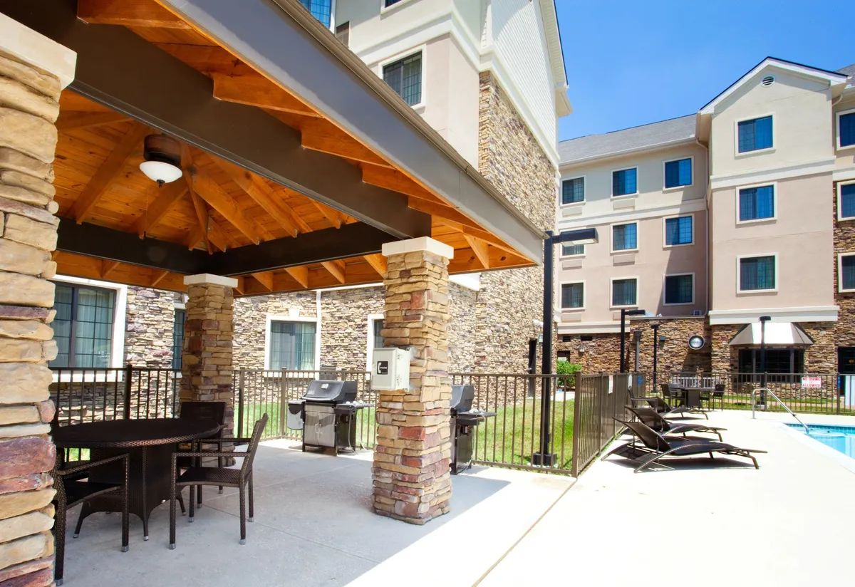 An inviting exterior space featuring an outdoor seating area, BBQ grill, swimming pool, and stone columns. People are gathered, barbecuing, and relaxing by the pool, suggesting a vibrant, sociable environment perfect for outdoor dining and evening gatherings.