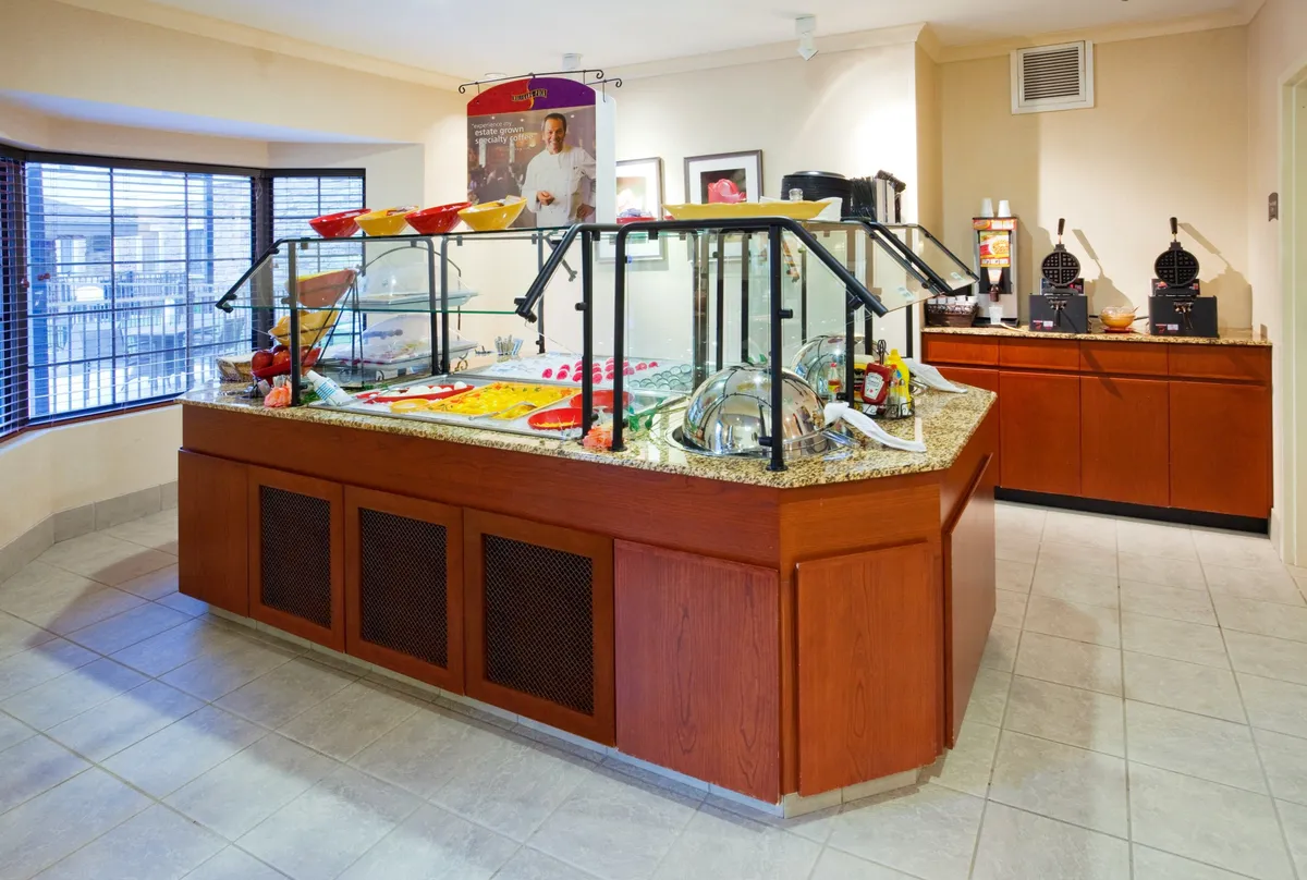 The scene shows a food display counter set up for a self-serve breakfast, with glass serving containers, decorative bowls, and a dedicated coffee station. Guests are seen enjoying the meal in a casual and welcoming atmosphere, emphasizing both convenience and social interaction.